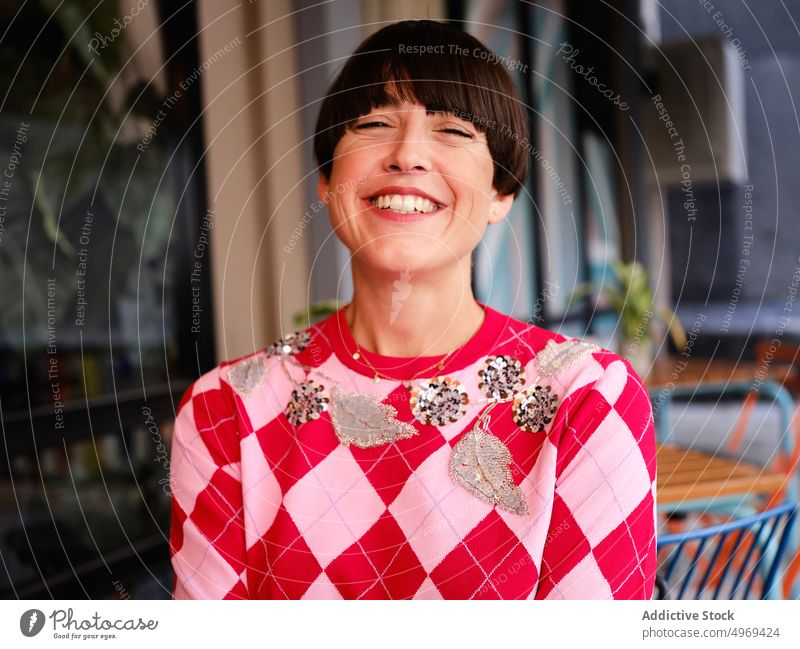 Positiv gestimmte Frau mit strahlendem Lächeln auf der Terrasse eines Sommercafés heiter trendy Café Inhalt stilvoll Frisur bob Stil Outfit modern Freude