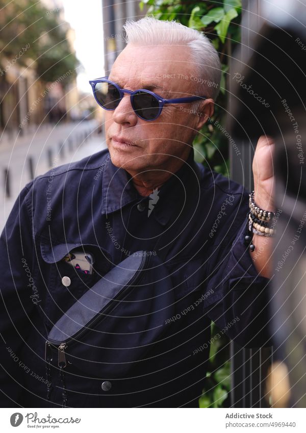 Modischer älterer Mann mit Gürteltasche auf der Straße Mode Senior trendy selbstbewusst gealtert Stil urban männlich Metall Zaun Hemd Sonnenbrille Großstadt