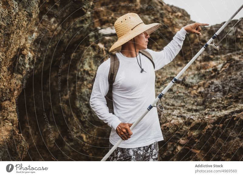 Anonymer Angler mit Angel in der Nähe einer Klippe Fischer Fischen Stab Küste Ufer Hobby Gerät Meeresufer Freizeit Berge u. Gebirge Felsen männlich Seeküste Hut