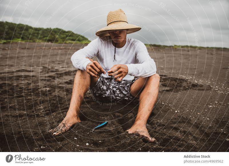 Unbekannter Fischer beim Stricken eines Fischerknotens Fischen stricken Knäuel Linie Küste Ufer Vorrat Gerät Hobby vorbereiten männlich Meeresufer Seeküste Hut