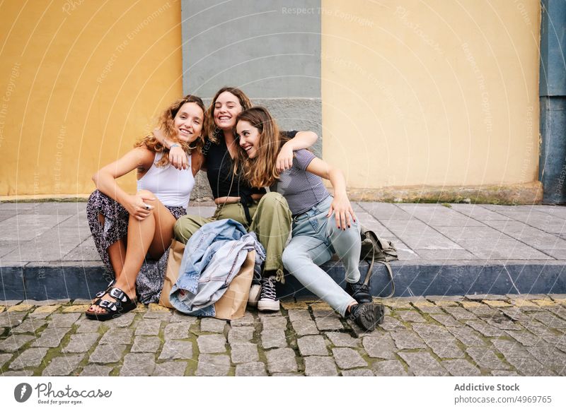 Frauen, die auf einer Asphaltkante auf der Straße sitzen und auf ihrem Smartphone surfen Browsen Sitzung benutzend Freundschaft heiter lässig Lächeln