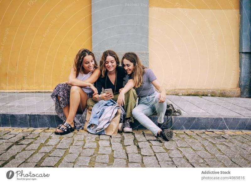Frauen browsen auf ihrem Smartphone in der Nähe einer bunten Wand Browsen Straße Sitzung benutzend Freundschaft heiter lässig Lächeln Zusammensein Mobile