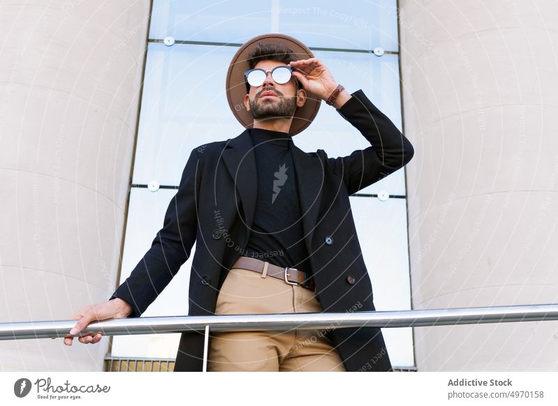 Maskuliner Mann mit trendiger Sonnenbrille hinter einem Geländer in der Stadt Mode Stil Individualität kreativ Design Zeitgenosse maskulin Hut Jacke Macho