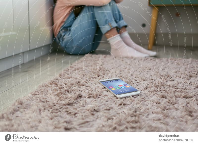 Auf dem Boden liegendes Mobiltelefon mit einem unerkennbar traurigen kleinen Mädchen, das dahinter sitzt Handy Lügen Stock unkenntlich Sitzen Schlafzimmer