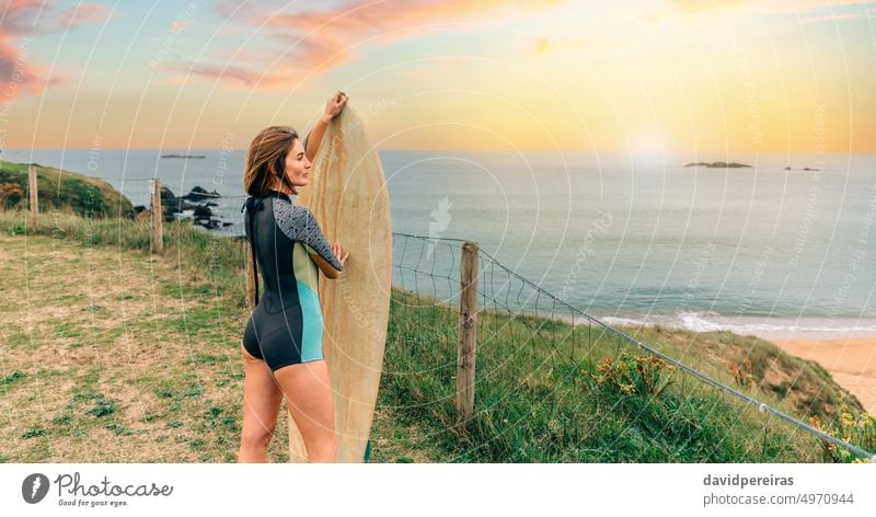 Surferin mit Neoprenanzug posiert mit Surfbrett mit Blick auf den Strand Frau jung posierend Textfreiraum Küste Sonnenuntergang Sonnenlicht Frühlingsanzug