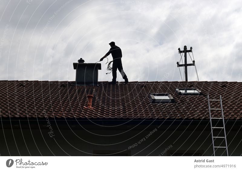 Ein Kaminkehrer säubert auf einem Hausdach den Kamin kaminkehrer schornsteinfeger haus gebäude mann hausdach säubern reinigen kehren bürste dachziegel oben höhe