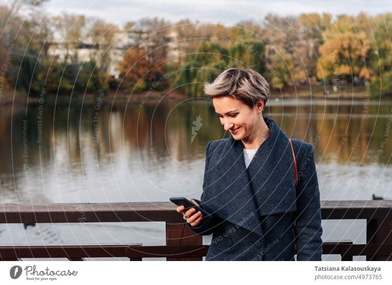 Eine Frau in einem Stadtpark am Fluss benutzt ein Smartphone Erwachsener Kurze Haare blond attraktiv Zelle Handy Fröhlichkeit Glück Freude Lächeln Lebensstile
