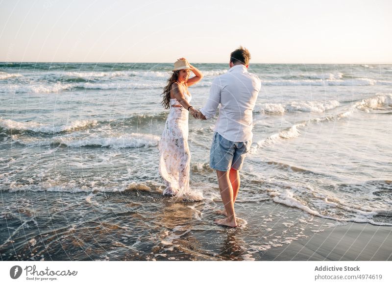 Zärtliches Paar, das bei Sonnenuntergang auf den Wellen des Meeres steht und die Hände hält Händchenhalten Stehen winken MEER Sommer verliebt sonnenbeschienen