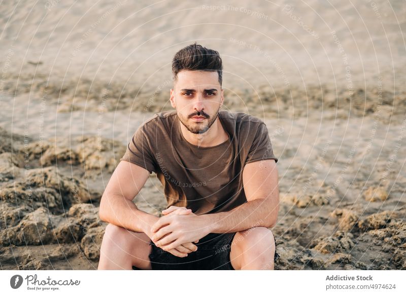Hübscher junger hispanischer Mann sitzt am Sandstrand und fasst sich an den Händen Strand nachdenklich beschaulich brünett aussruhen Falten Sitzen entspannend