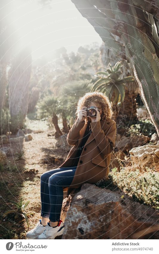 Porträt einer schönen lockigen blonden Frau mit einer Retro-Kamera, die auf einem Felsen sitzt Natur im Freien Accessoire Bekleidung Fotokamera Mantel