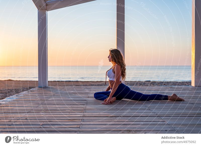 Frau übt Yoga in Taubenhaltung am Meeresufer meditieren üben Sonnenaufgang Namaste ruhig gestikulieren hölzern Spazierweg Seeküste Asana Pose