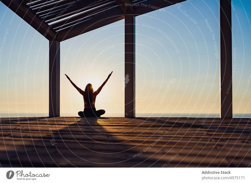 Ruhige Frau in Lotus-Pose am Meer bei Sonnenaufgang Yoga Seeküste Silhouette meditieren Morgen Harmonie Asana Terrasse hölzern padmasana schlank Morgendämmerung