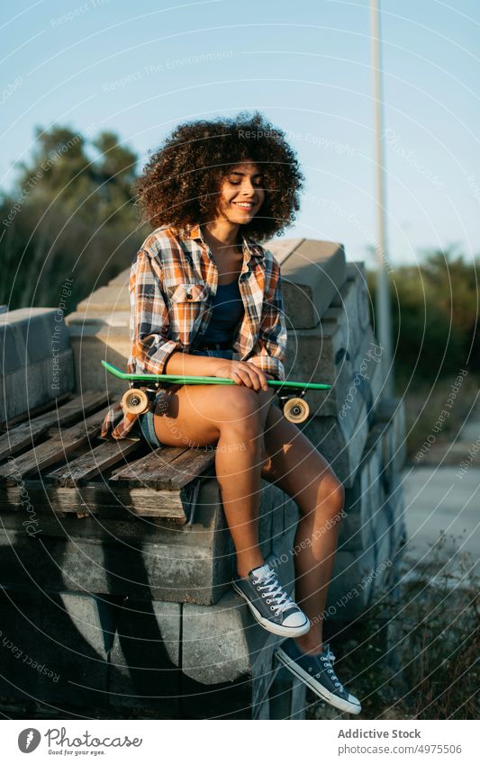 Afroamerikanische Frau mit Penny Board sitzt auf Steinen auf der Straße Lochrasterplatte Sommer Skater cool tausendjährig Afro-Look trendy ruhig Hipster sonnig