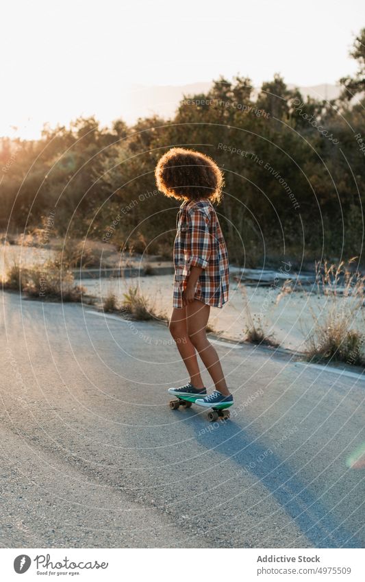 Ethnische Frau reitet Penny Board Lochrasterplatte Mitfahrgelegenheit Sonnenuntergang Straße Sommer Hipster Skater Afro-Look urban ethnisch trendy ruhig