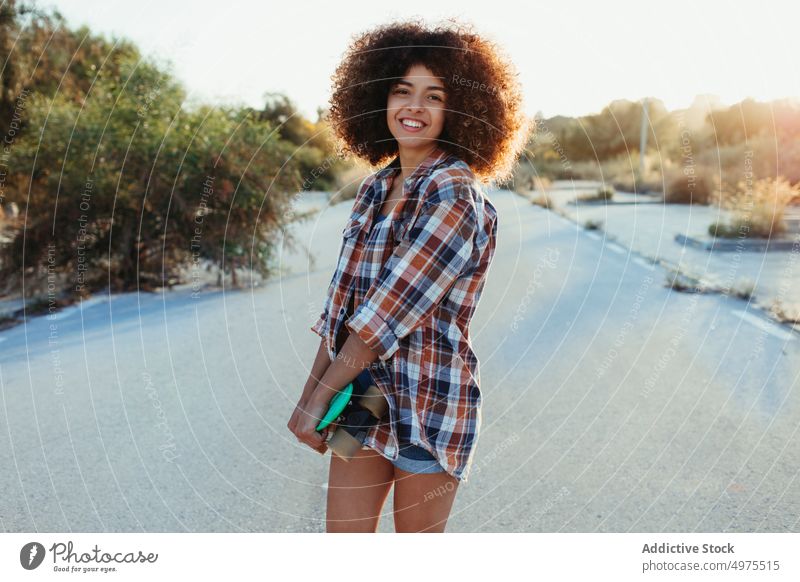 Fröhliche ethnische Frau mit Penny Board Lochrasterplatte Straße Hipster Sommer Skater Afro-Look Sonnenuntergang heiter tausendjährig Afroamerikaner schwarz