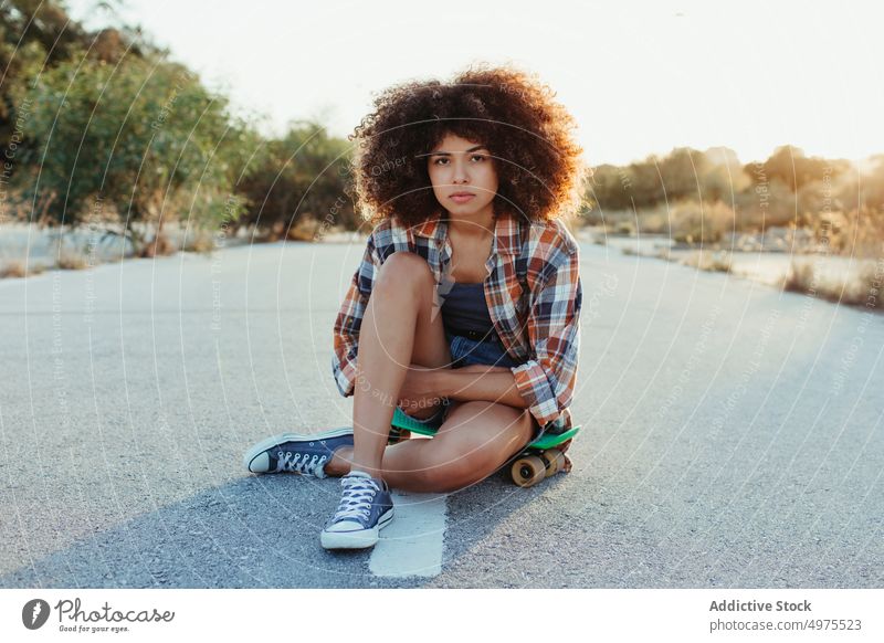 Afroamerikanische Frau sitzt auf einem Penny Board auf der asphaltierten Straße Lochrasterplatte Sommer Skater cool tausendjährig Afro-Look Asphalt trendy ruhig