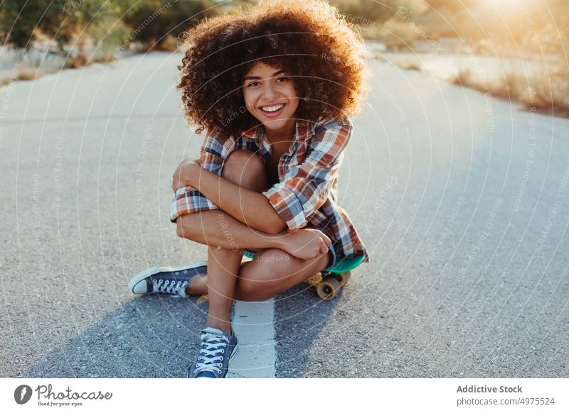 Entzückende afroamerikanische Frau, die auf einem Penny Board auf der Asphaltstraße sitzt Lochrasterplatte Sommer Skater Straße cool tausendjährig Afro-Look