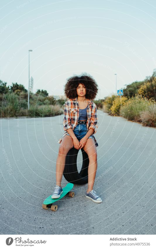 Entzückende afroamerikanische Frau auf Reifen sitzend mit Penny Board auf der Asphaltstraße Lochrasterplatte Sommer Skater Straße cool tausendjährig Afro-Look