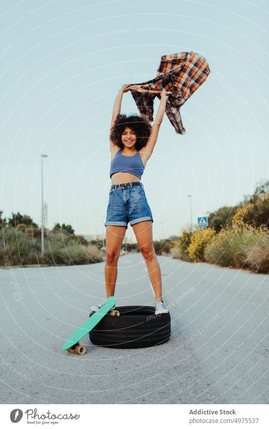 Entzückende afroamerikanische Frau auf Reifen sitzend mit Penny Board auf der Asphaltstraße Lochrasterplatte Sommer Skater Straße cool tausendjährig Afro-Look