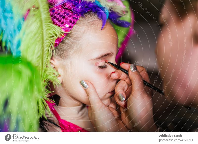 Crop Mutter Anwendung Festival Make-up auf Tochter Gesicht Karneval vorbereiten Verkehrsflugzeug bewerben heimwärts farbenfroh Feder Kopfbedeckung Mädchen Frau