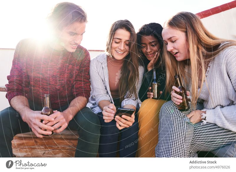 Fröhliche Freunde mit Bier und Smartphone auf dem Sofa auf der Terrasse benutzend Inhalt Party heiter Schnaps Zusammensein Gerät Apparatur zuschauen Glück