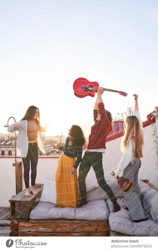 Begeisterte multiethnische Freunde spielen Gitarre und springen auf dem Terrassensofa Dachterrasse aufgeregt Musik Instrument Spaß haben Party fröhlich