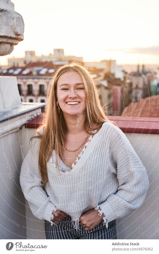 Lächelnde junge Frau auf der Terrasse stehend heiter Zahnfarbenes Lächeln Stimmung Glück Dachterrasse Dämmerung Stadt expressiv aufgeregt lässig Inhalt Reling