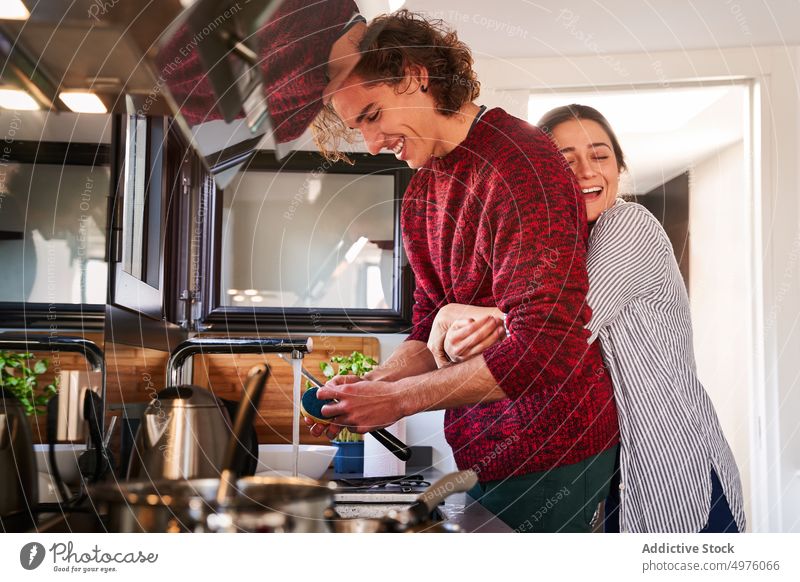 Freudige Frau umarmt ihren Freund beim gemeinsamen Kochen in der Küche Paar Umarmung Freude Waschen Waschbecken Zahnfarbenes Lächeln Umarmen Zusammensein