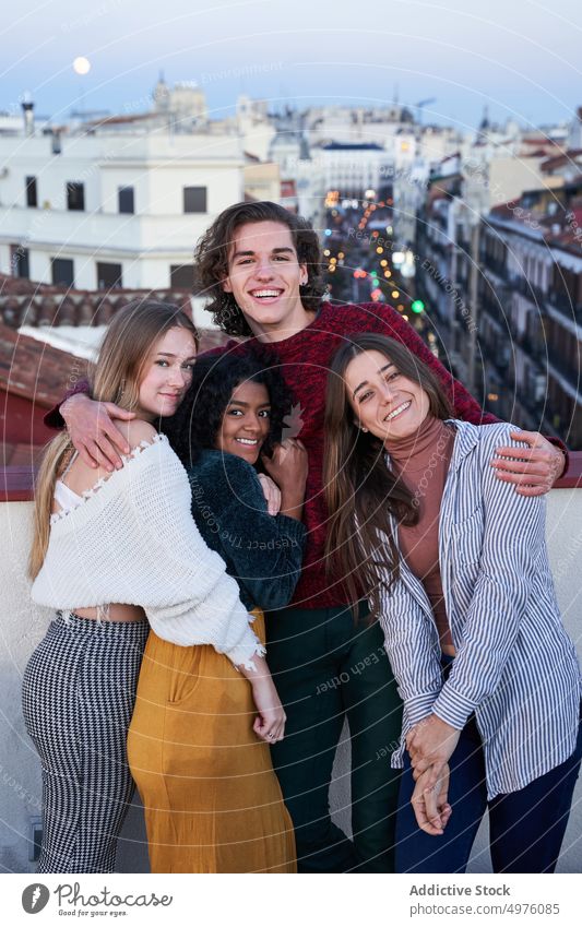 Lächelnde multirassische Freunde, die sich umarmen und in der Dämmerung auf einem Dach sitzen Umarmung Dachterrasse Zahnfarbenes Lächeln heiter Zusammensein