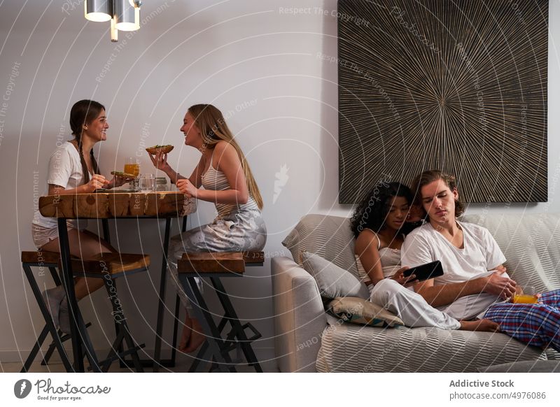 Fröhliche Frauen beim Abendessen in der Nähe von Mitbewohnern mit Smartphone auf dem Sofa Paar Freund Wohnzimmer benutzend Zusammensein heiter Apparatur dine