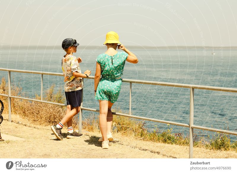 Zwei Kinder in Sommerkleidung unterhalten sich, am Geländer vor dem Meer Sonne T-Shirt Kleid Hut Gespräch Austausch Wärme Unterhaltung lässig leger vertieft
