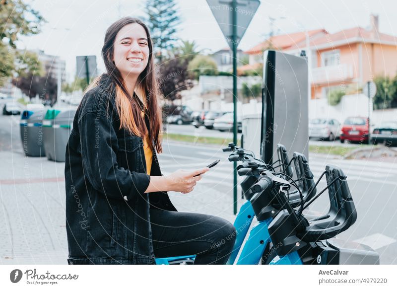 Junge Frau überprüft lächelnd ihr Telefon, um ein Fahrrad zu entsperren und auszuleihen. Sie prüft, ob ein Fahrrad verfügbar ist und wie der Batteriestatus ist. Elektrisches Verkehrskonzept. Erneuerbare Ressourcen - Bewegung in der Stadt