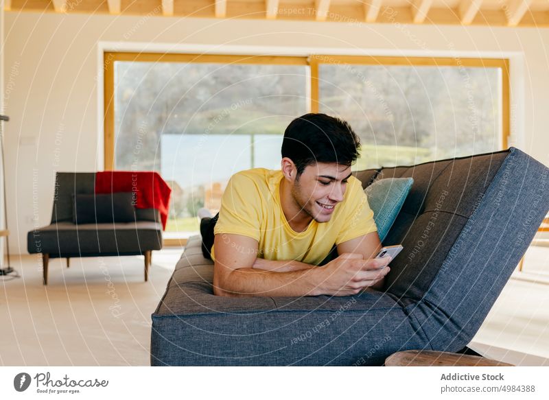 Junger lächelnder Mann auf Sofa liegend mit Smartphone im Zimmer Raum Lügen hispanisch Typ Kanapee Handy Fenster Lächeln benutzend Glück groß jung gemütlich