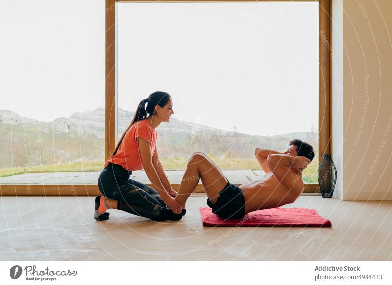 Glückliche Frau, die einem Mann beim Training im Zimmer am Fenster hilft Paar Hilfe Raum Sport sich aufsetzen Übungen ohne Hemd Bein jung schlank groß