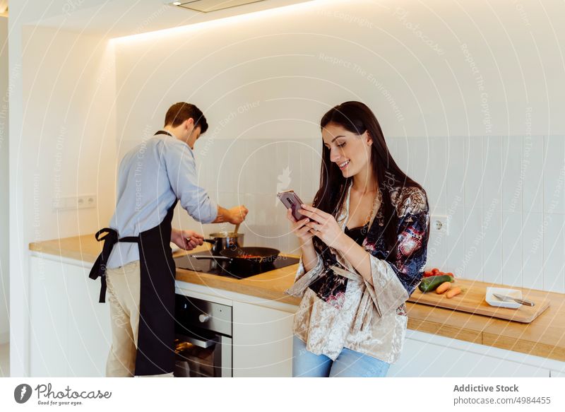 Glückliche Frau mit Smartphone in der Nähe von Mann bereitet Essen in der Küche Paar Lebensmittel Vorbereitung hispanisch Bratpfanne Topf Elektroherd Schürze
