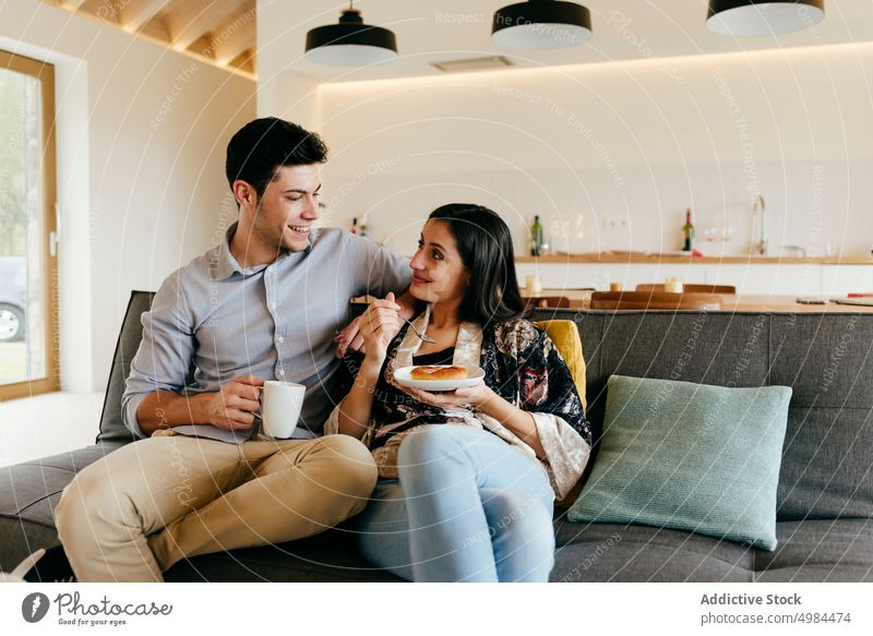 Glückliches Paar mit Tasse und Teller auf Sofa im Zimmer Raum hispanisch Küche Becher Löffel Kanapee heiter Lächeln Frau Mann jung attraktiv Getränk trinken