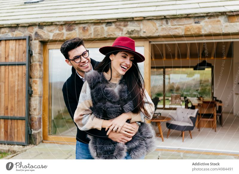 Glückliches Paar, das sich in der Nähe eines modernen Hauses umarmt umarmend hispanisch Frau Mann Brille Rücken Hut Cottage Lächeln heiter jung attraktiv