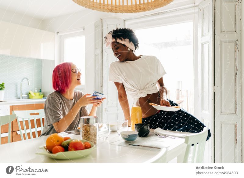Fröhliche junge Frau mit Smartphone in der Nähe des kochenden schwarzen Freundes Frauen benutzend Küche heimwärts Morgen Frühstück Lächeln Koch vielfältig