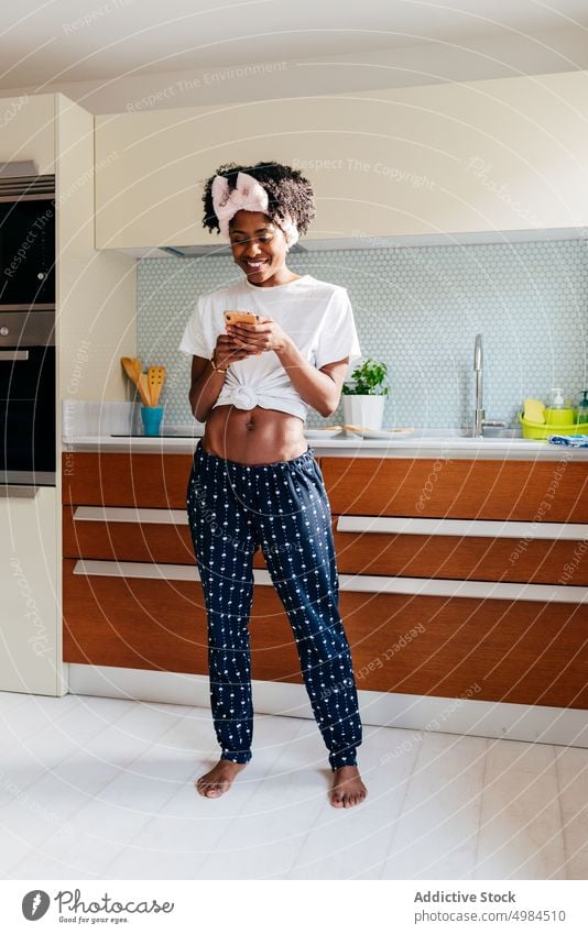 Fröhliche schwarze Frau benutzt Smartphone in der Küche benutzend Lächeln Appartement modern Stil ethnisch gemütlich Lifestyle heimwärts sich[Akk] entspannen
