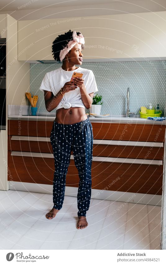 Fröhliche schwarze Frau benutzt Smartphone in der Küche benutzend Lächeln Appartement modern Stil ethnisch gemütlich Lifestyle heimwärts sich[Akk] entspannen