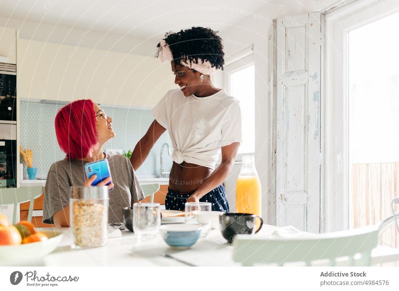 Fröhliche junge Frau mit Smartphone in der Nähe des kochenden schwarzen Freundes Frauen benutzend Küche heimwärts Morgen Frühstück Lächeln Koch vielfältig