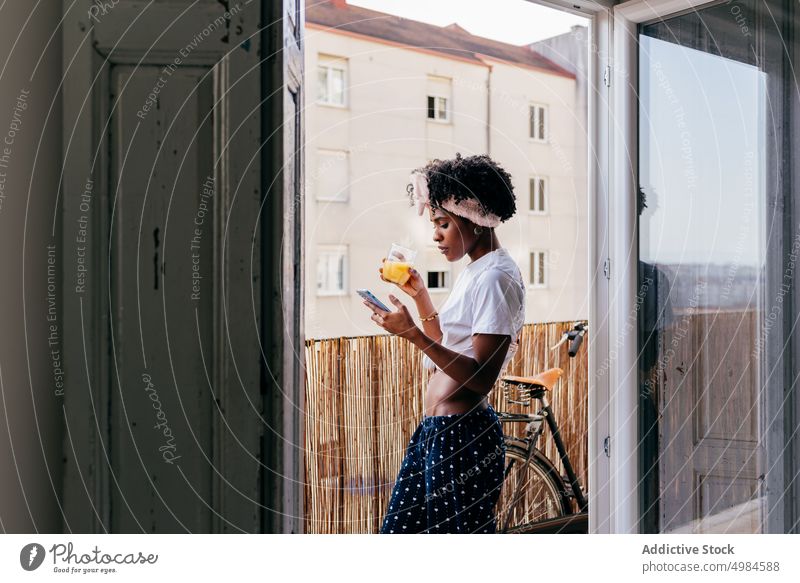 Gesunde schwarze Frau, die auf dem Balkon ihr Smartphone durchsucht Browsen zuschauend Saft Lächeln ethnisch Frühstück modern Morgen trinken jung Getränk Tasse