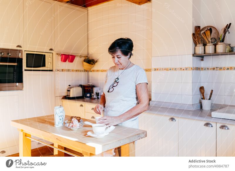 Ältere Frau bereitet Frühstück in der Küche vor heimwärts vorbereiten Lebensmittel Morgen hölzern Innenbereich Möbel Mahlzeit Hausfrau reif Licht beige Keramik