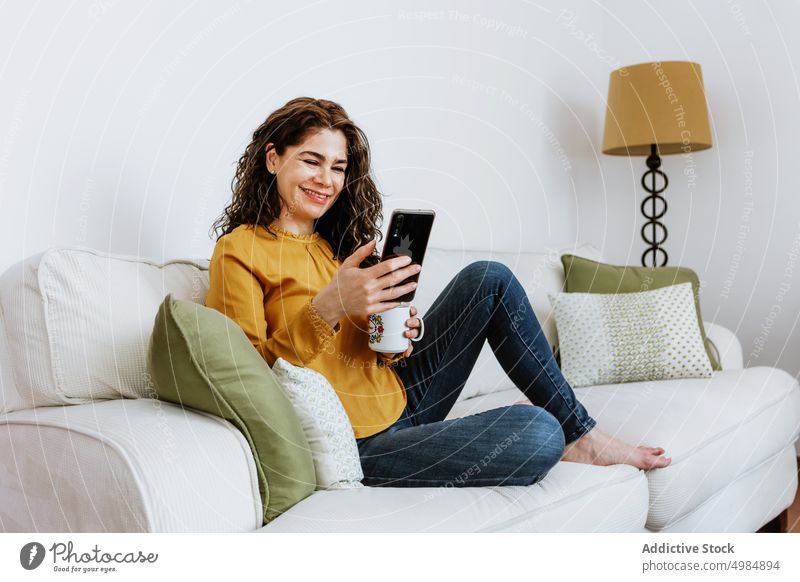 Lächelnde Frau, die sich zu Hause per Videoanruf über ihr Smartphone unterhält positiv benutzend Browsen heiter Tasse Kaffee Tee Zahnfarbenes Lächeln