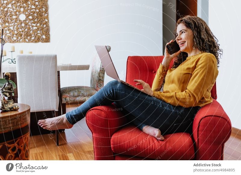 Glückliche Frau, die mit ihrem Smartphone spricht, während sie im Wohnzimmer auf ihrem Laptop surft positiv freiberuflich Telefonanruf reden Kommunizieren