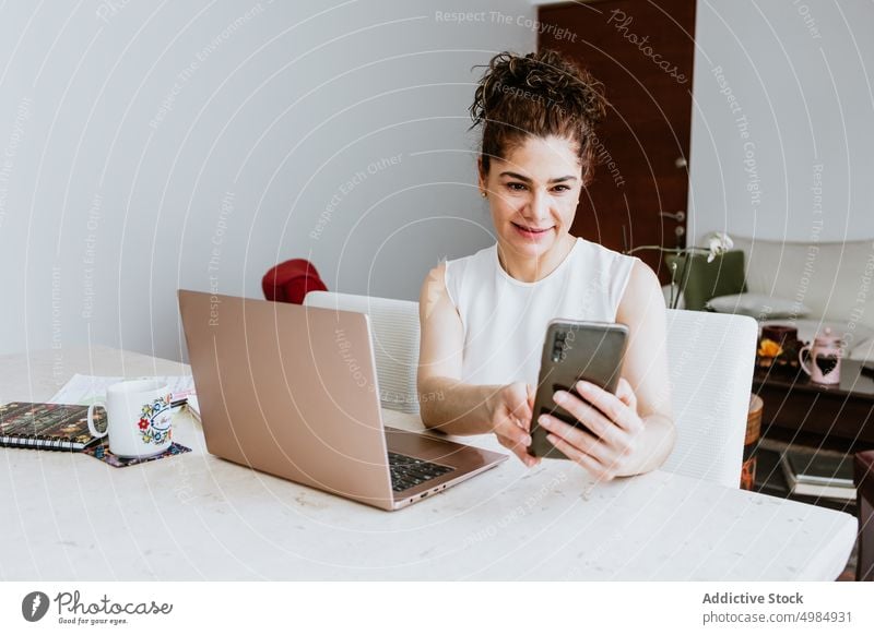 Lächelnde Frau surft auf ihrem Smartphone, während sie mit ihrem Laptop am Tisch sitzt freiberuflich positiv Videoanruf Arbeit Beruf Unternehmer heiter