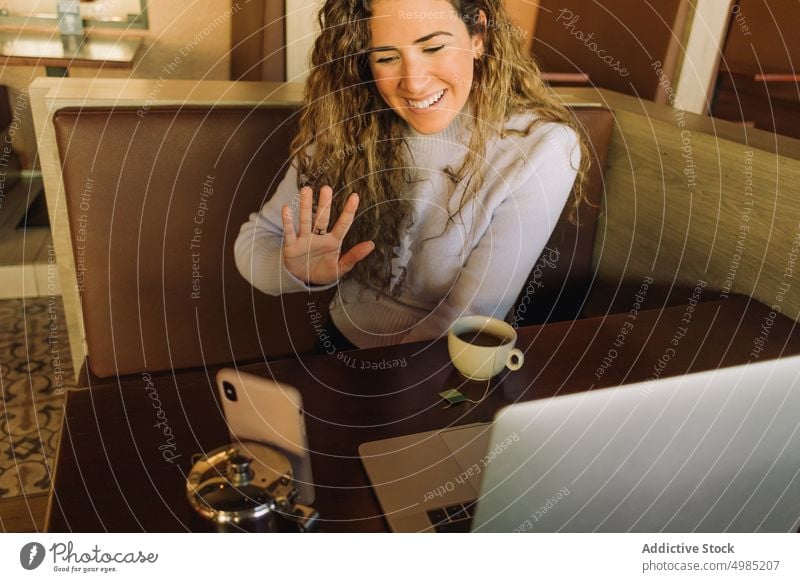 Fröhliche Frau mit winkender Hand bei einem Videoanruf in einem Cafe freiberuflich Wellenhand Tee heiter Lächeln Laptop Smartphone Café reden Gerät Hallo