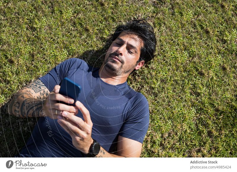 Müder ethnischer Sportler, der auf grünem Rasen auf seinem Smartphone surft Browsen müde Pause Läufer Lügen Gras Surfen online männlich hispanisch Mann