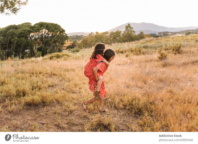Lächelndes Mädchen trägt Schwester auf dem Rücken in einem Feld Huckepack Sommer Natur Mitfahrgelegenheit führen Urlaub Glück sorgenfrei Freund Spaß Wiese rot