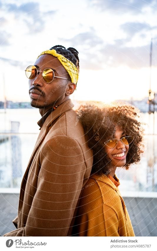 Stilvolles schwarzes Paar in Mänteln auf der Stadtpromenade Pier trendy urban farbenfroh Herbst Lachen ethnisch Afroamerikaner Mode Afro-Look Rastalocken Frisur